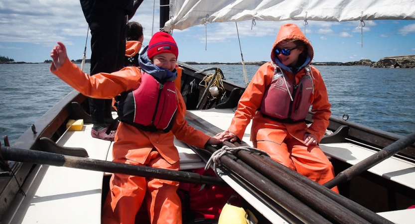 Two people wearing orange drysuits and lifejackets sit in a sailboat on calm water